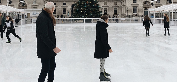 a father and daughter in winter attire iceskating on an ice rink with a christmas tree in the background