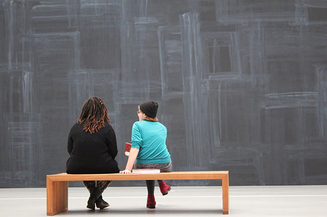 two females sitting on a bench facing a blank chalkboard wall